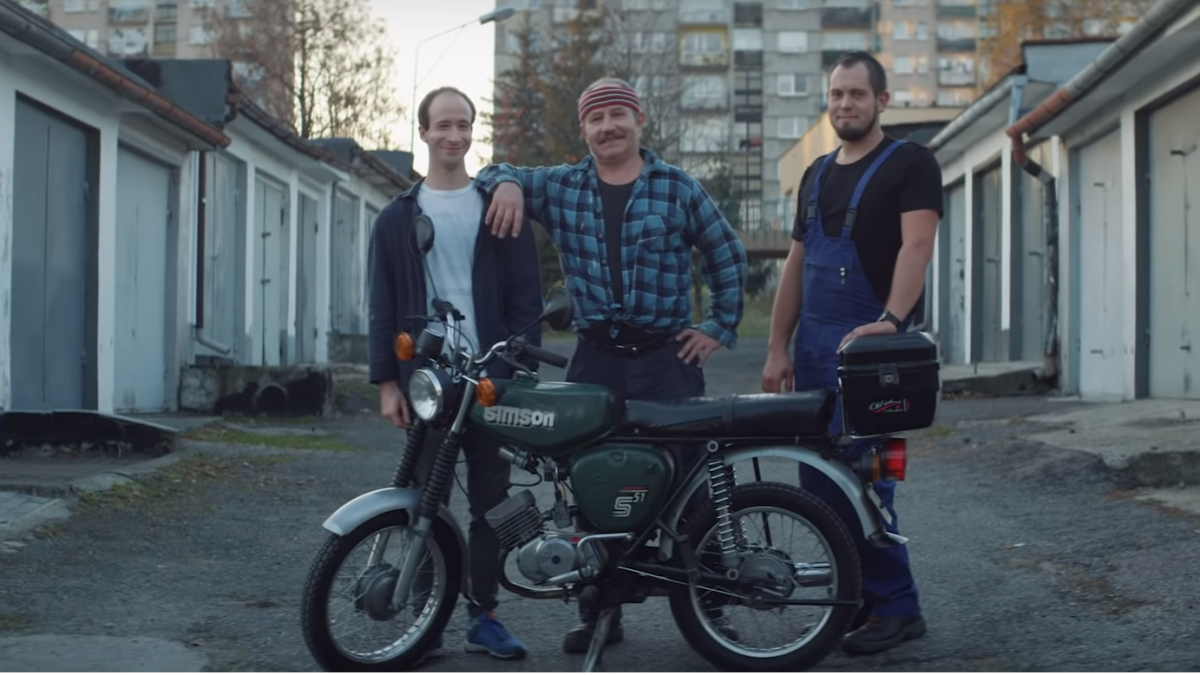 Trzech mężczyzn stojących obok siebie, przed nimi motor, w tle bloki mieszkalne