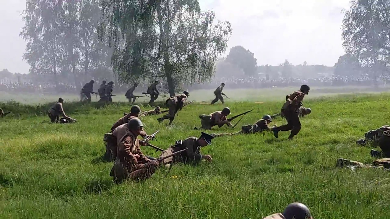 Żołnierze biorący udział w inscenizacji biegnący po trawie z drzewem w tle