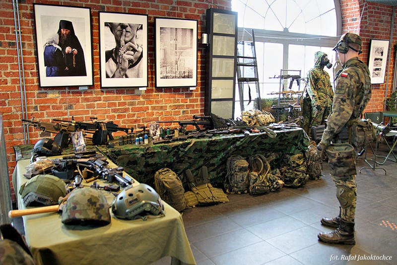 Na stołach leży wyposażenie żołnierzy jak hełmy czy imitacje broni, po prawej strony stoi mężczyzna ubrany w strój żołnierza