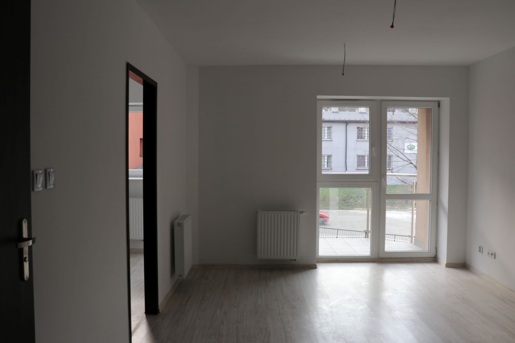 Pomieszczenie z pomalowanymi na biało ścianami z oknem balkonowym, po lewej stronie drzwi do innego pomieszczenia
