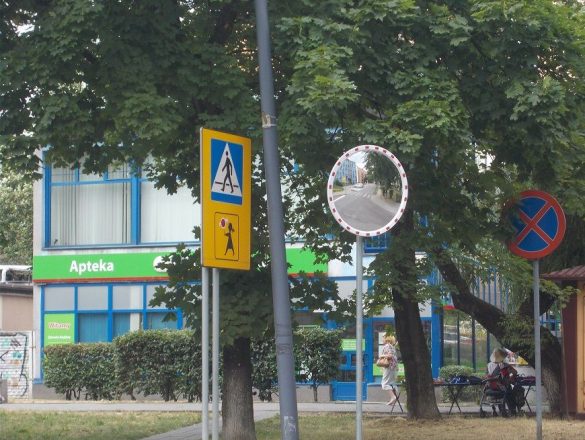 Znaki drogowy obok lustro drogowe, w tle drzewa i apteka