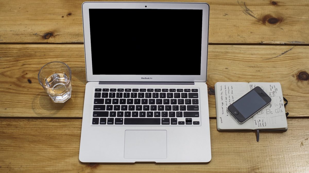 Laptop obok którego stoi szklanka wody oraz leżą telefon i notatnik