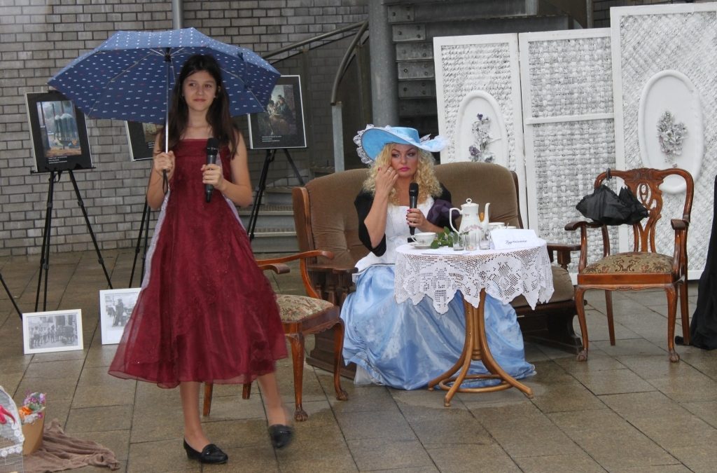 Dziewczynka w czerwonej sukni i pod parasolem oraz siedząca kobieta w kapeluszu podczas inscenizacjia noweli Dobra Pani Elizy Orzeszkowej