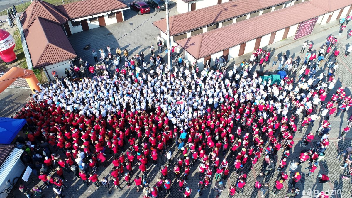 Zdjęcie z góry, uczestnicy biegu w koszulkach białych i czerwonych tworzą flagę stojąc przed startem na targowisku miejskim