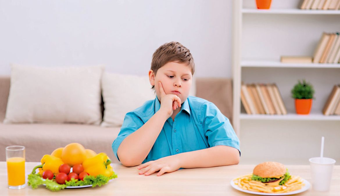 Młody chłopiec siedzący przy stole przed dwoma posiłkami: talerzem warzyw oraz hamburgerem. Na twarzy chłopca widoczny jest grymas zastanowienia. Swój wzrok kieruje w stronę hamburgera.