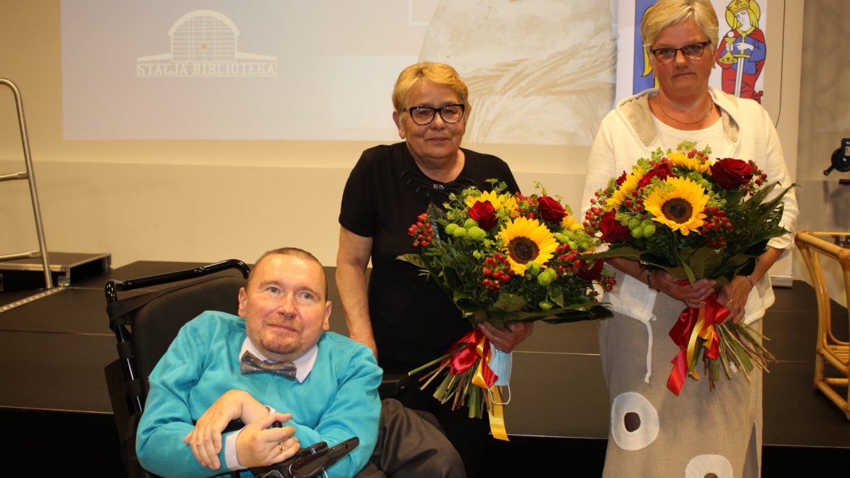 Mężczyzna na wózku inwalidzkim oraz dwie kobiety z bukietami kwiatów, w tle prezentacja wyświetlona na ekranie.