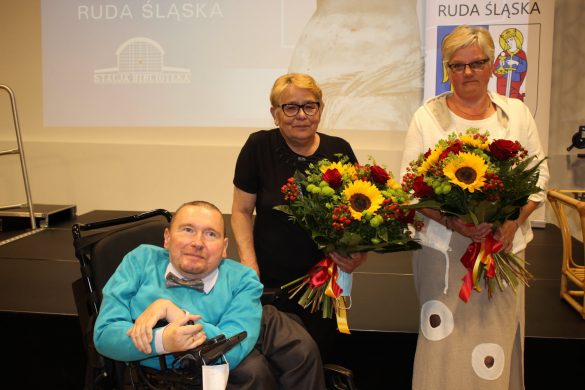 Mężczyzna na wózku inwalidzkim oraz dwie kobiety z bukietami kwiatów, w tle prezentacja wyświetlona na ekranie.