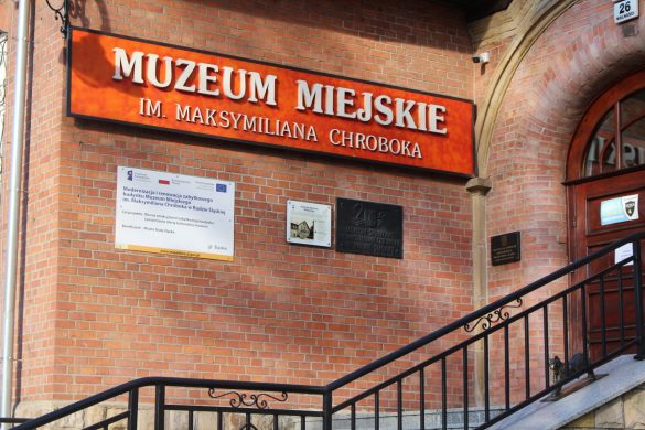 Fragment budynku z cegły z napisem "Muzeum Miejskie im. Maksymiliana Chroboka" i tablicami informacyjnymi, schody z poręczą prowadzące do brązowych drzwi