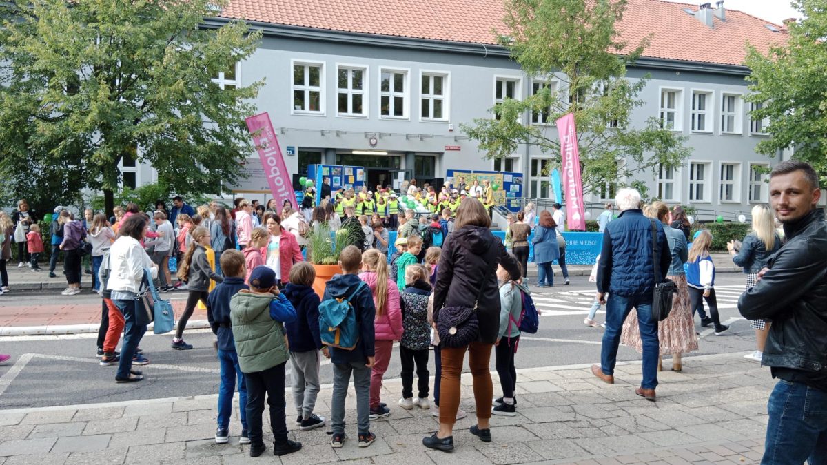 Grupa dzieci i rodziców stoi na chodniku przed szkołą