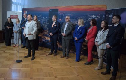 Obraz przedstawia grupę dziesięciu osób na drewnianej podłodze, z nocną panoramą miasta i napisami "INNOWACYJNY KONCEPT POMOCY" oraz "fundacja wolne miejsce" w tle. Po lewej stronie stoi kobieta w ciemnym garniturze, obok kobieta w szarym kostiumie i mężczyzna w ciemnym garniturze z czerwonym krawatem. Dalej mężczyzna w jasnym blezerze, kobieta w jasnej bluzce i ciemnej spódnicy oraz mężczyzna w ciemnym garniturze z pomarańczowym krawatem. W środku mężczyzna w szarym garniturze, po jego prawej kobieta w niebieskim i kobieta w czerwonym kostiumie. Po prawej dwie kobiety: w jasnej bluzce i beżowych spodniach oraz w jasnej bluzce i ciemnych spodniach. Na końcu po prawej mężczyzna w ciemnym garniturze. Osoby patrzą w różnych kierunkach, niektóre przemawiają. Przed nimi mikrofon na statywie. Oświetlenie jest jasne, w tle widać okno z zasłonami.
