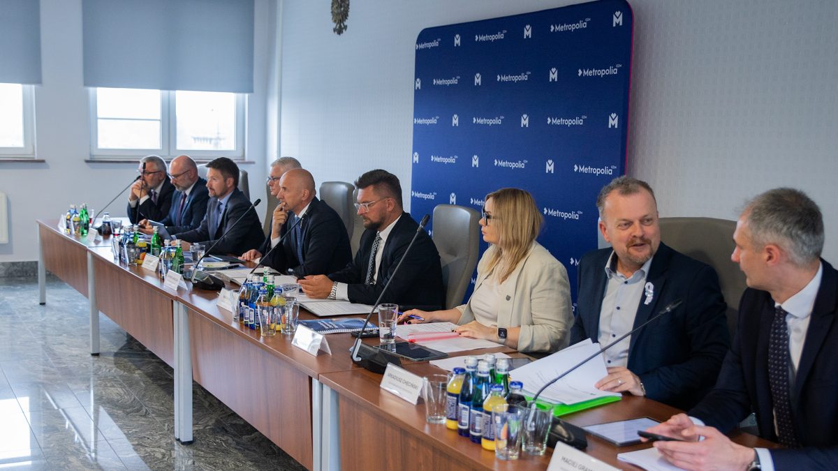 Na zdjęciu widać grupę około dziesięciu osób, siedzących przy długim, drewnianym stole konferencyjnym w dobrze oświetlonym pomieszczeniu. Większość z nich to mężczyźni w średnim wieku, ubrani w garnitury i koszule. Po prawej stronie stołu siedzi jedna kobieta w jasnej marynarce, która aktywnie uczestniczy w rozmowie. Na stole znajdują się butelki z wodą, szklanki, dokumenty i foldery. W tle widać niebieską ścianę z logo 