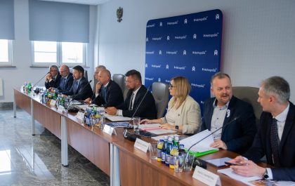 Na zdjęciu widać grupę około dziesięciu osób, siedzących przy długim, drewnianym stole konferencyjnym w dobrze oświetlonym pomieszczeniu. Większość z nich to mężczyźni w średnim wieku, ubrani w garnitury i koszule. Po prawej stronie stołu siedzi jedna kobieta w jasnej marynarce, która aktywnie uczestniczy w rozmowie. Na stole znajdują się butelki z wodą, szklanki, dokumenty i foldery. W tle widać niebieską ścianę z logo "Metropolia GZM" powtórzonym wielokrotnie. Pomieszczenie wydaje się być salą konferencyjną lub biurem.