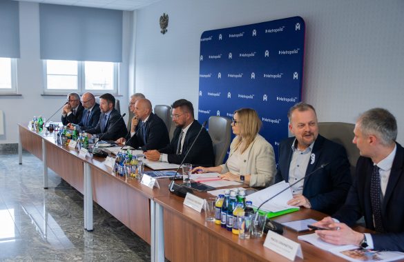 Na zdjęciu widać grupę około dziesięciu osób, siedzących przy długim, drewnianym stole konferencyjnym w dobrze oświetlonym pomieszczeniu. Większość z nich to mężczyźni w średnim wieku, ubrani w garnitury i koszule. Po prawej stronie stołu siedzi jedna kobieta w jasnej marynarce, która aktywnie uczestniczy w rozmowie. Na stole znajdują się butelki z wodą, szklanki, dokumenty i foldery. W tle widać niebieską ścianę z logo "Metropolia GZM" powtórzonym wielokrotnie. Pomieszczenie wydaje się być salą konferencyjną lub biurem.