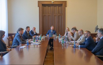 Zdjęcie przedstawia spotkanie w długiej sali konferencyjnej. Na środku stołu, zwrócony przodem do obiektywu, siedzi mężczyzna w garniturze. Po obu stronach stołu siedzą inne osoby, niektóre patrzą na mówcę, inne na laptopy. Na stole znajdują się butelki z wodą i szklanki. W tle widać drewniane drzwi i jasne ściany.