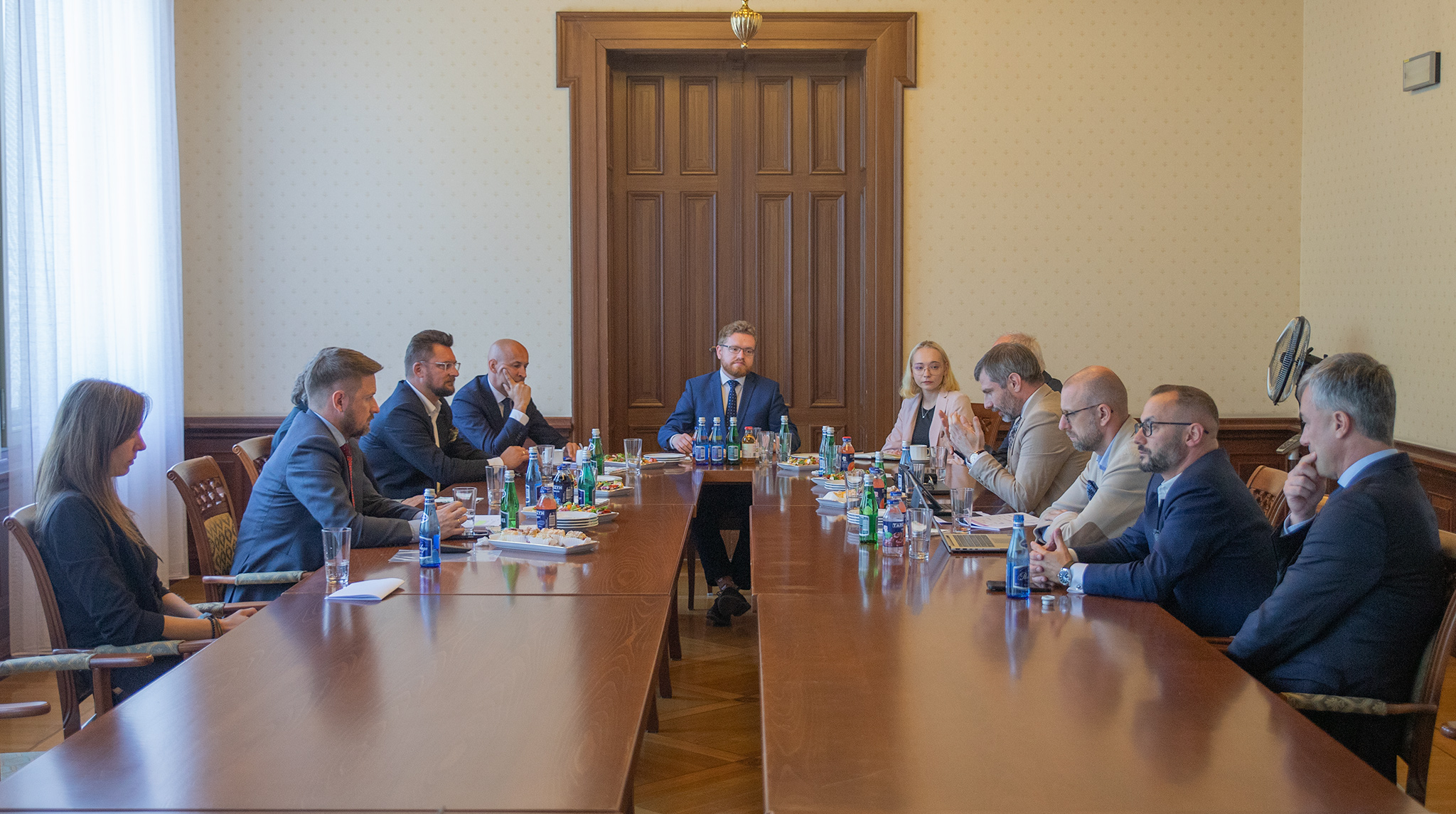 Zdjęcie przedstawia spotkanie w długiej sali konferencyjnej. Na środku stołu, zwrócony przodem do obiektywu, siedzi mężczyzna w garniturze. Po obu stronach stołu siedzą inne osoby, niektóre patrzą na mówcę, inne na laptopy. Na stole znajdują się butelki z wodą i szklanki. W tle widać drewniane drzwi i jasne ściany.