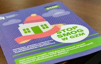 Obraz przedstawia fioletową ulotkę kampanii "STOP SMOG w GZM". Centralnie umieszczony jest rysunek domu z szalikiem, otoczonego chmurami. Tekst na ulotce informuje o poprawie jakości powietrza w Zagłębiowskiej Metropolii i programie finansowania ocieplenia domów.