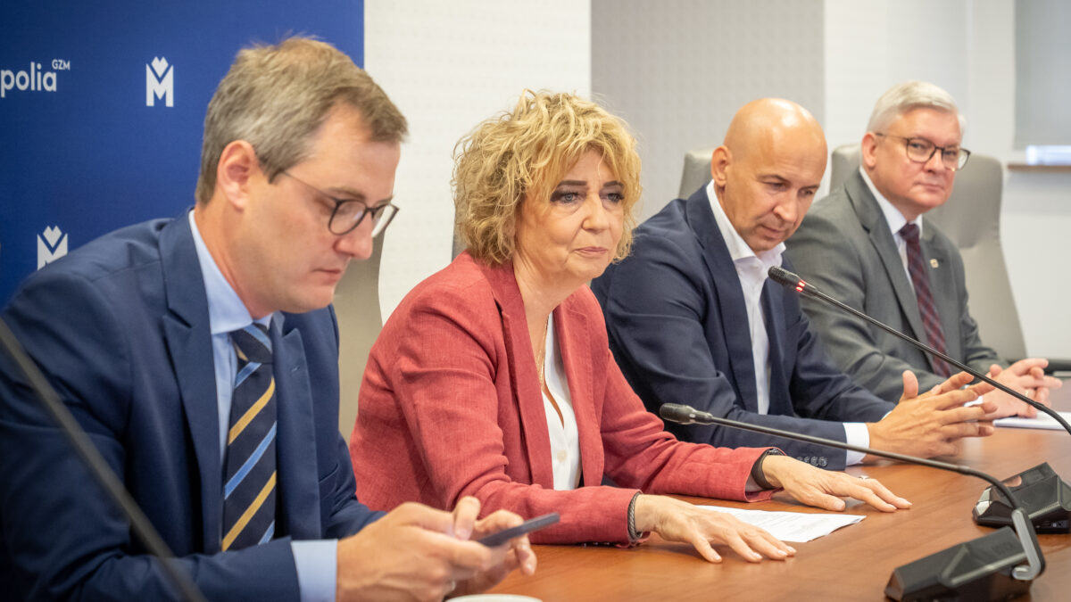 Na zdjęciu czterech ludzi siedzi przy drewnianym stole. Młody mężczyzna w okularach patrzy w telefon. Obok niego kobieta w czerwonej marynarce spogląda przed siebie, a przed nią leżą dokumenty i smartfon. Obok niej łysy mężczyzna w ciemnym garniturze patrzy w dół. Dalej siedzi starszy mężczyzna w okularach. Przed każdą z osób znajduje się mikrofon.