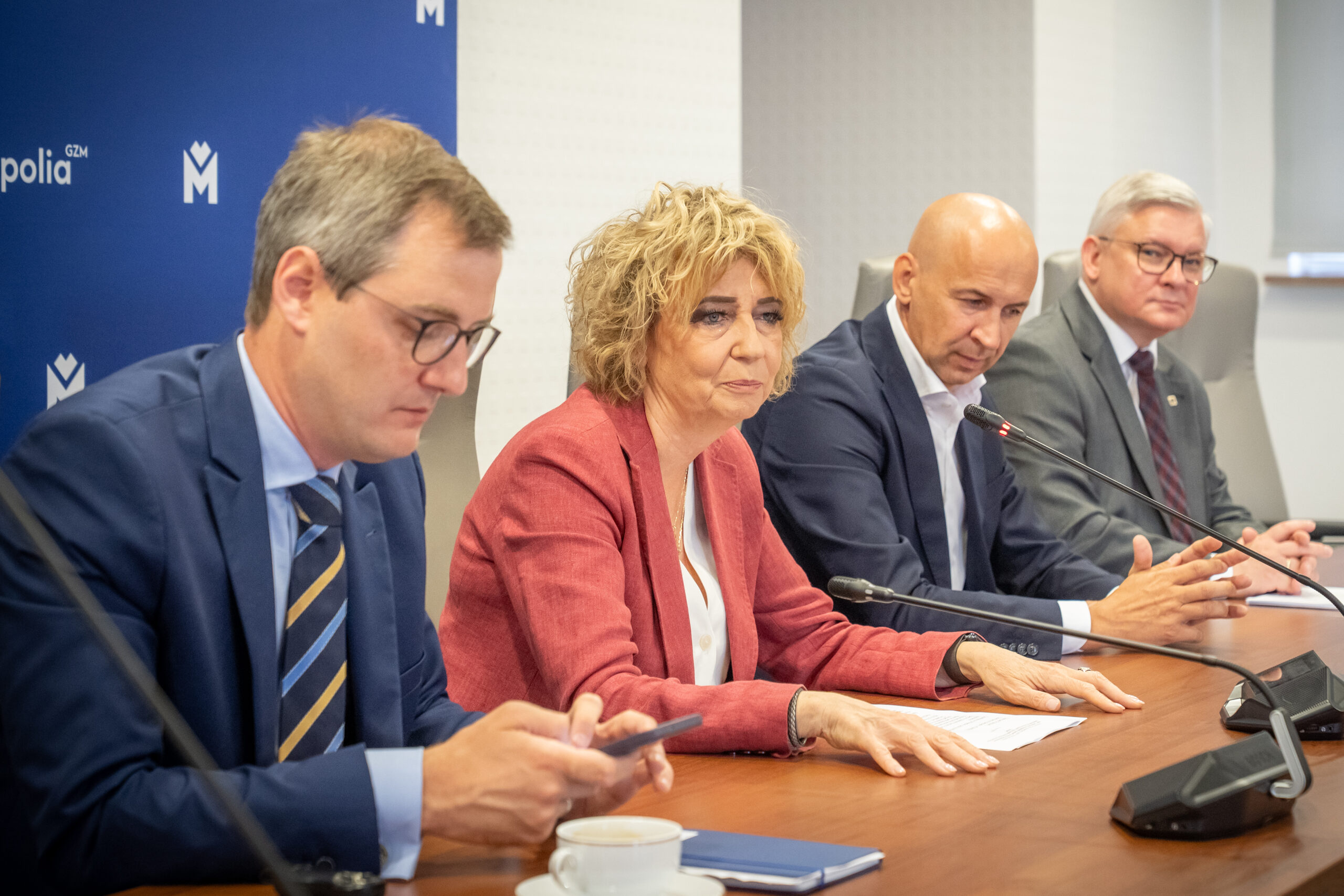 Na zdjęciu czterech ludzi siedzi przy drewnianym stole. Młody mężczyzna w okularach patrzy w telefon. Obok niego kobieta w czerwonej marynarce spogląda przed siebie, a przed nią leżą dokumenty i smartfon. Obok niej łysy mężczyzna w ciemnym garniturze patrzy w dół. Dalej siedzi starszy mężczyzna w okularach. Przed każdą z osób znajduje się mikrofon.