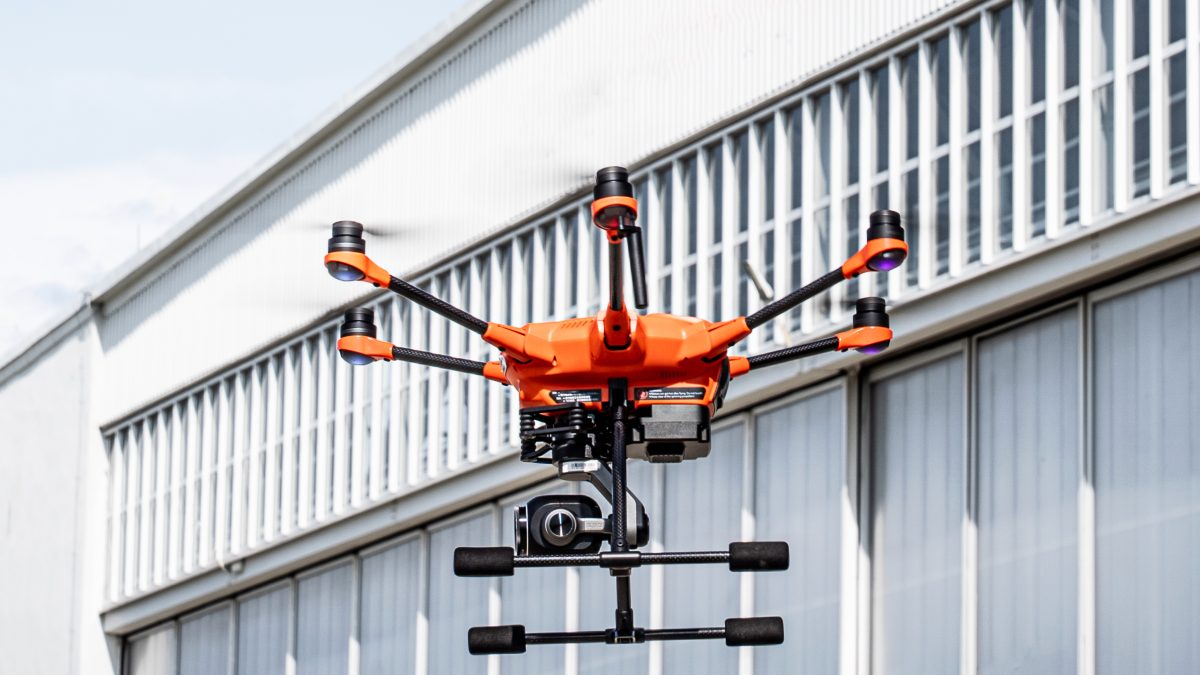 Na zdjęciu widoczny jest pomarańczowo-czarny dron z sześcioma ramionami i kamerą, unoszący się w powietrzu na tle jasnego, panelowego budynku. Pogoda jest słoneczna.