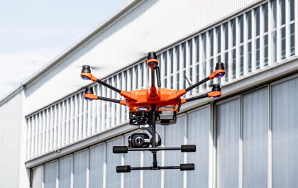 Na zdjęciu widoczny jest pomarańczowo-czarny dron z sześcioma ramionami i kamerą, unoszący się w powietrzu na tle jasnego, panelowego budynku. Pogoda jest słoneczna.