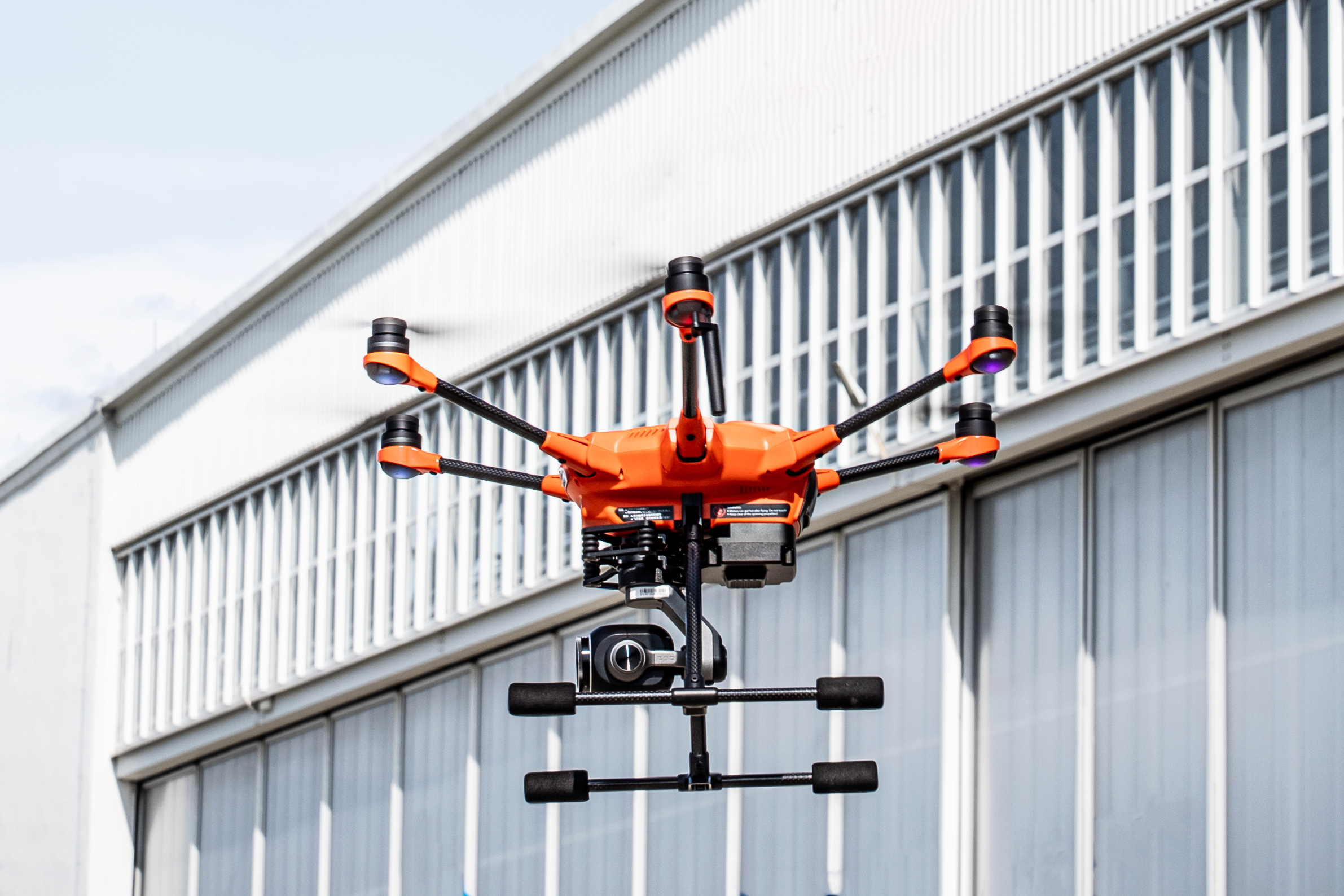 Na zdjęciu widoczny jest pomarańczowo-czarny dron z sześcioma ramionami i kamerą, unoszący się w powietrzu na tle jasnego, panelowego budynku. Pogoda jest słoneczna.