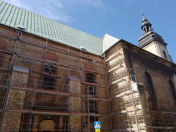 Zdjęcie przedstawia fragment Kościoła pw. św. Wojciecha, ukazując jego boczną ścianę i część wieży. Budynek jest częściowo pokryty rusztowaniami, co może wskazywać na trwające prace konserwatorskie lub remontowe. Mur kościoła wykonany jest z cegły i kamienia, co świadczy o jego historycznym charakterze. Dach kościoła ma zielonkawy odcień. W tle widać fragment błękitnego nieba.