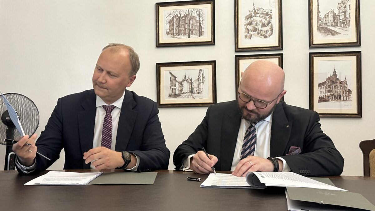 Na zdjęciu widać dwóch mężczyzn siedzących przy stole, podpisujących dokumenty. Jeden z nich, w okularach i z brodą, pisze, podczas gdy drugi obserwuje go, trzymając w ręku papier. W tle wiszą oprawione grafiki przedstawiające budynki.