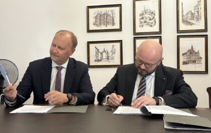 Na zdjęciu widać dwóch mężczyzn siedzących przy stole, podpisujących dokumenty. Jeden z nich, w okularach i z brodą, pisze, podczas gdy drugi obserwuje go, trzymając w ręku papier. W tle wiszą oprawione grafiki przedstawiające budynki.