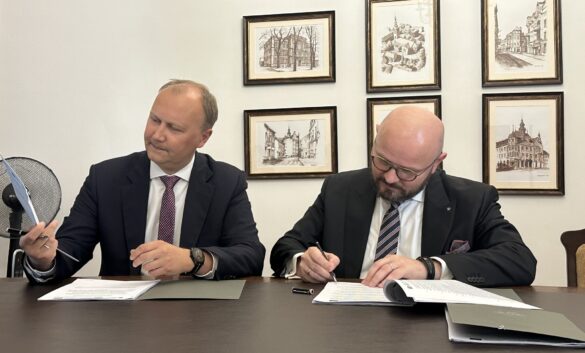Na zdjęciu widać dwóch mężczyzn siedzących przy stole, podpisujących dokumenty. Jeden z nich, w okularach i z brodą, pisze, podczas gdy drugi obserwuje go, trzymając w ręku papier. W tle wiszą oprawione grafiki przedstawiające budynki.