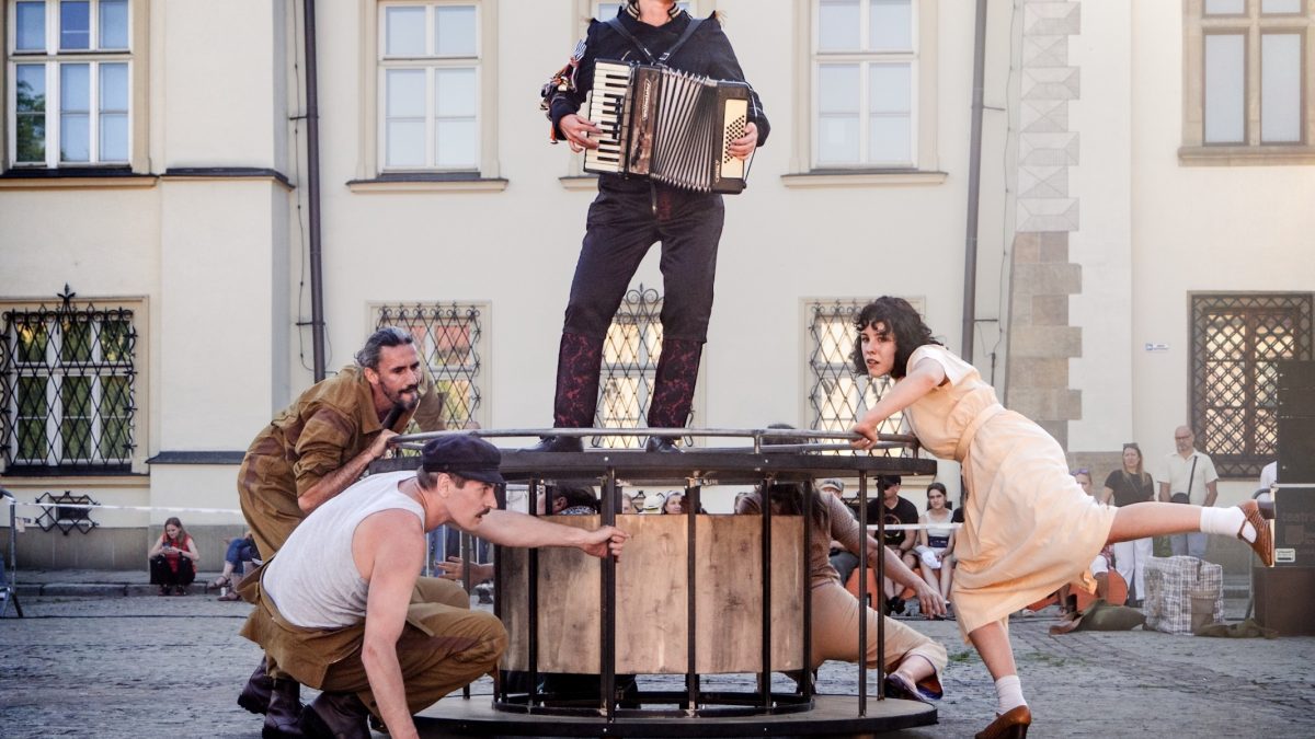 Zdjęcie przedstawia scenę z występu na świeżym powietrzu, prawdopodobnie teatralnego. W centrum kadru znajduje się konstrukcja przypominająca okrągły, drewniany stół lub scenę, na której stoi osoba grająca na akordeonie. Wokół tej konstrukcji znajdują się trzy inne osoby: dwie kucają, wchodząc w interakcję z elementami konstrukcji, a jedna stoi obok, opierając się o nią. Wszyscy aktorzy mają na sobie stroje stylizowane, co sugeruje charakterystyczny występ. W tle widoczne są zabytkowe budynki oraz grupa obserwujących widzów.