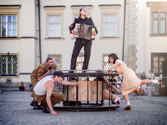 Zdjęcie przedstawia scenę z występu na świeżym powietrzu, prawdopodobnie teatralnego. W centrum kadru znajduje się konstrukcja przypominająca okrągły, drewniany stół lub scenę, na której stoi osoba grająca na akordeonie. Wokół tej konstrukcji znajdują się trzy inne osoby: dwie kucają, wchodząc w interakcję z elementami konstrukcji, a jedna stoi obok, opierając się o nią. Wszyscy aktorzy mają na sobie stroje stylizowane, co sugeruje charakterystyczny występ. W tle widoczne są zabytkowe budynki oraz grupa obserwujących widzów.