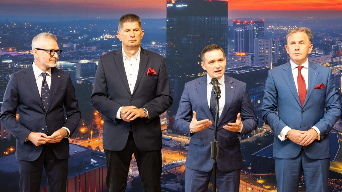 Na zdjęciu czterech mężczyzn w garniturach stoi przed tle panoramy miasta nocą. Jeden z mężczyzn, stojący w centrum, przemawia do mikrofonu, gestykulując. Pozostali trzej stoją obok niego, obserwując.