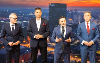 Na zdjęciu czterech mężczyzn w garniturach stoi przed tle panoramy miasta nocą. Jeden z mężczyzn, stojący w centrum, przemawia do mikrofonu, gestykulując. Pozostali trzej stoją obok niego, obserwując.