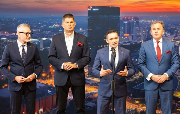 Na zdjęciu czterech mężczyzn w garniturach stoi przed tle panoramy miasta nocą. Jeden z mężczyzn, stojący w centrum, przemawia do mikrofonu, gestykulując. Pozostali trzej stoją obok niego, obserwując.