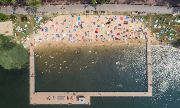 Zdjęcie z lotu ptaka przedstawia plażę i kąpielisko Stawiki w Sosnowcu w upalny dzień. Na piaszczystym brzegu leżakuje wielu ludzi, rozłożyli ręczniki i parasole. W wodzie, w wydzielonej strefie, pływają kąpiący się. Obok kąpieliska widać fragment otwartego akwenu.