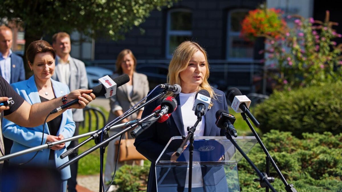 Na zdjęciu widać blondynkę w granatowym żakiecie i białej bluzce, która przemawia do kilku mikrofonów z logo różnych mediów. Za nią stoi grupa osób. Scena rozgrywa się na zewnątrz, w otoczeniu zieleni.