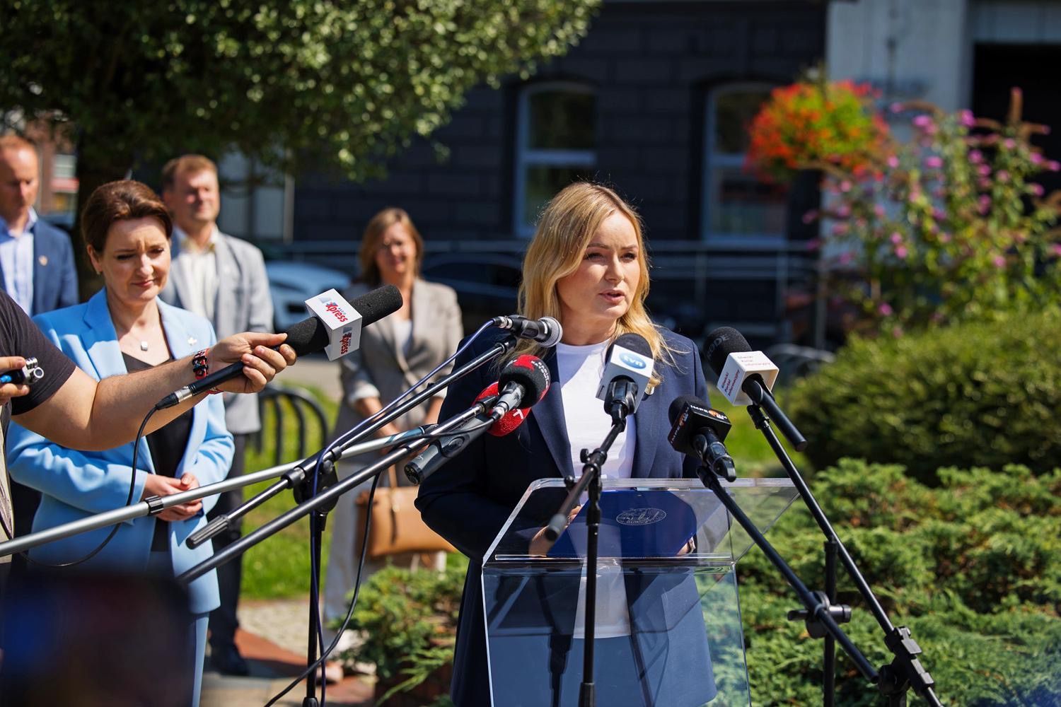 Na zdjęciu widać blondynkę w granatowym żakiecie i białej bluzce, która przemawia do kilku mikrofonów z logo różnych mediów. Za nią stoi grupa osób. Scena rozgrywa się na zewnątrz, w otoczeniu zieleni.