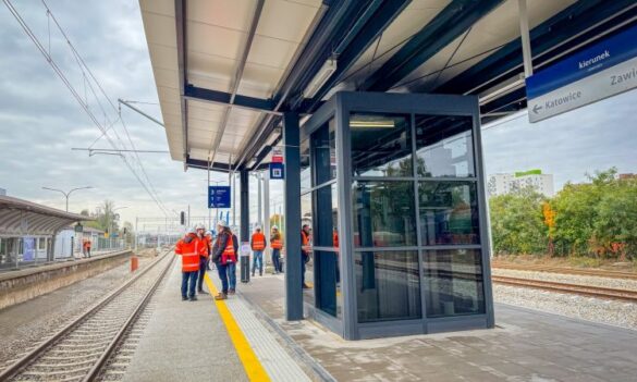 Nowy peron kolejowy, po remoncie, z nowoczesną wiatą i przeszkloną poczekalnią. Na peronie stoją pracownicy w pomarańczowych kamizelkach i kaskach. Widoczny jest znak kierunkowy "Katowice $\rightarrow$" i "Zawiercie". Stan torowiska świadczy, że prace są na ukończeniu lub zakończone.