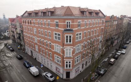 Widok z góry na narożną, odrestaurowaną kamienicę z czerwonej cegły. Budynek stoi na skrzyżowaniu miejskich ulic z zaparkowanymi autami.