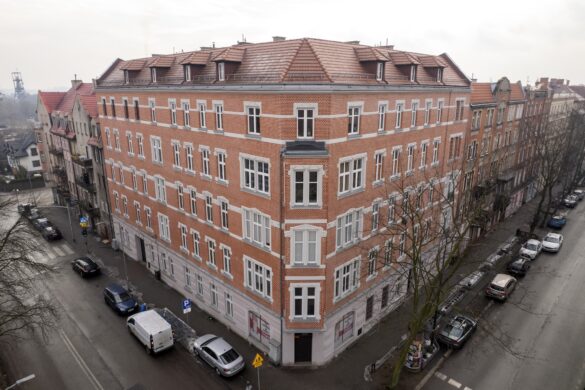 Widok z góry na narożną, odrestaurowaną kamienicę z czerwonej cegły. Budynek stoi na skrzyżowaniu miejskich ulic z zaparkowanymi autami.
