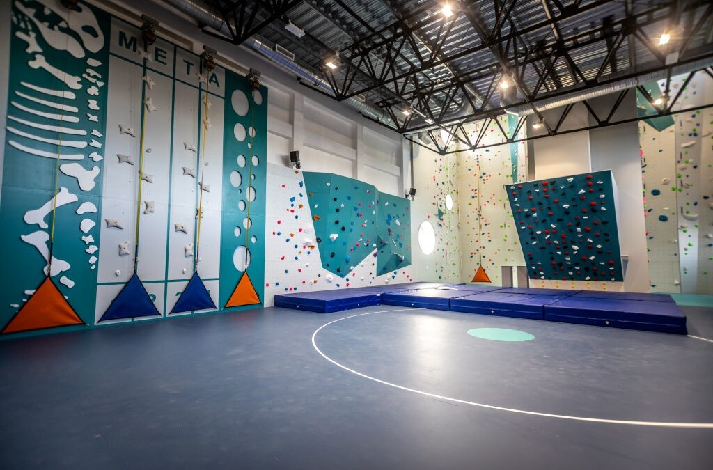 Zdjęcie przedstawia nowoczesną, kolorową ściankę wspinaczkową w hali sportowej. Widoczne są różnorodne trasy, materace asekuracyjne oraz wysoki sufit.
