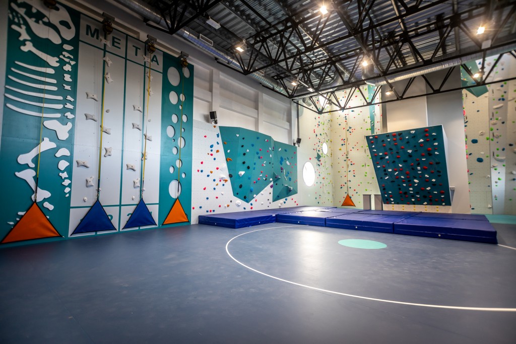 Zdjęcie przedstawia nowoczesną, kolorową ściankę wspinaczkową w hali sportowej. Widoczne są różnorodne trasy, materace asekuracyjne oraz wysoki sufit.