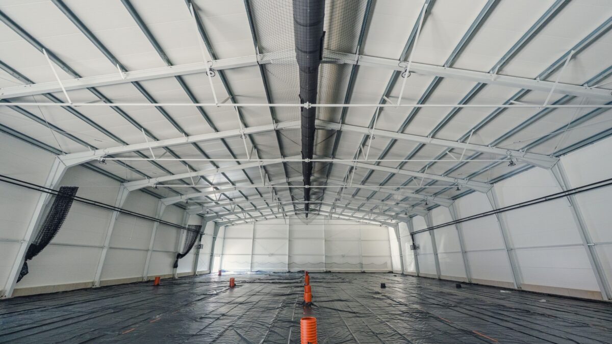 Wnętrze dużej, pustej hali o białej konstrukcji. Podłoga przykryta jest czarną folią, a na środku stoją pomarańczowe pachołki.