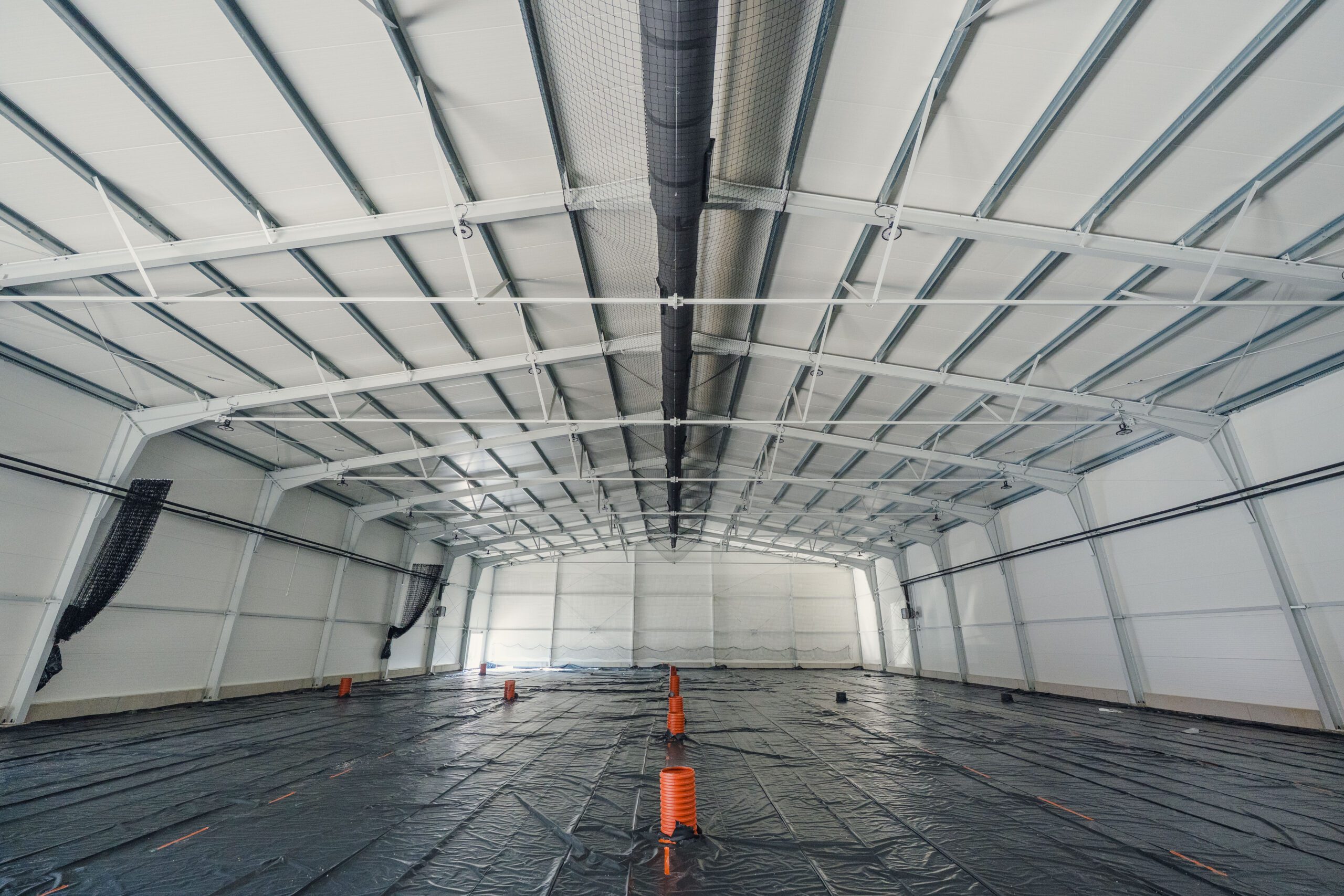Wnętrze dużej, pustej hali o białej konstrukcji. Podłoga przykryta jest czarną folią, a na środku stoją pomarańczowe pachołki.