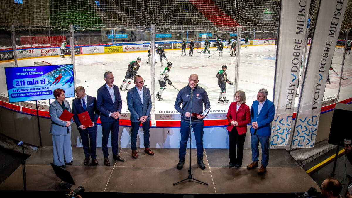 Konferencja na lodowisku w Tychach. Grupa osób w garniturach stoi przed ekranem z napisem 211 mln zł na budowę lodowisk. W tle trenują hokeiści.
