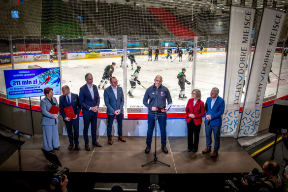 Konferencja na lodowisku w Tychach. Grupa osób w garniturach stoi przed ekranem z napisem 211 mln zł na budowę lodowisk. W tle trenują hokeiści.
