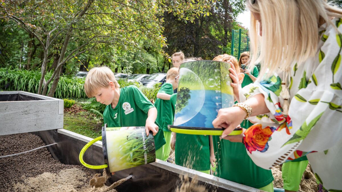 Dzieci w zielonych strojach wsypują piasek z wiader do dużej skrzyni.