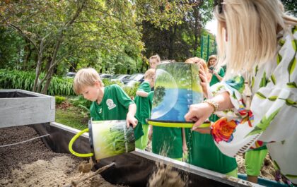 Dzieci w zielonych strojach wsypują piasek z wiader do dużej skrzyni.