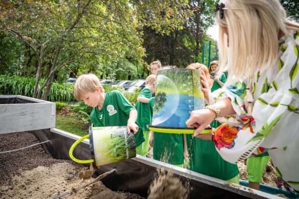 Dzieci w zielonych strojach wsypują piasek z wiader do dużej skrzyni.