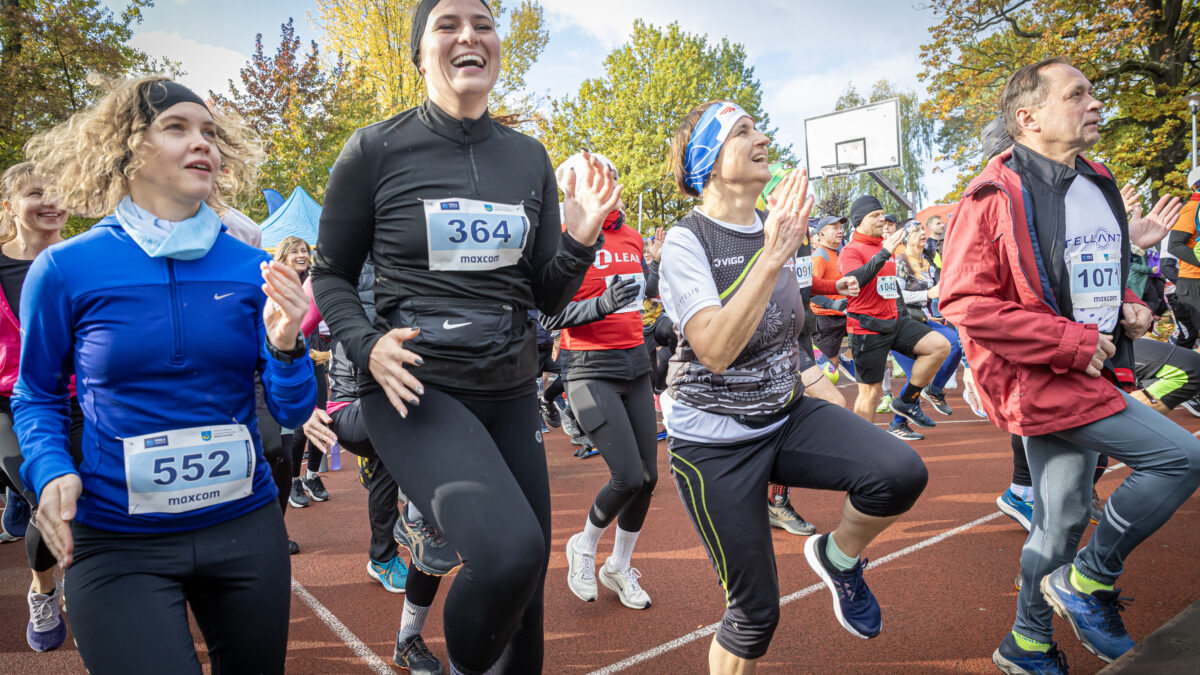 Gromada uśmiechniętych biegaczy na czerwonej bieżni podczas rozgrzewki. Ludzie w kolorowych strojach sportowych z numerami startowymi radośnie podskakują.